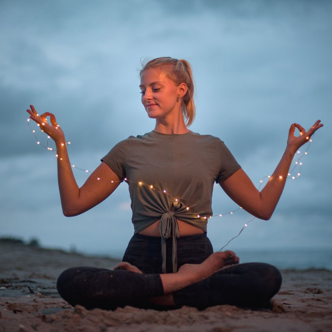 Corey Heyman meditating on the beach at dusk with string lights wrapped around her hands